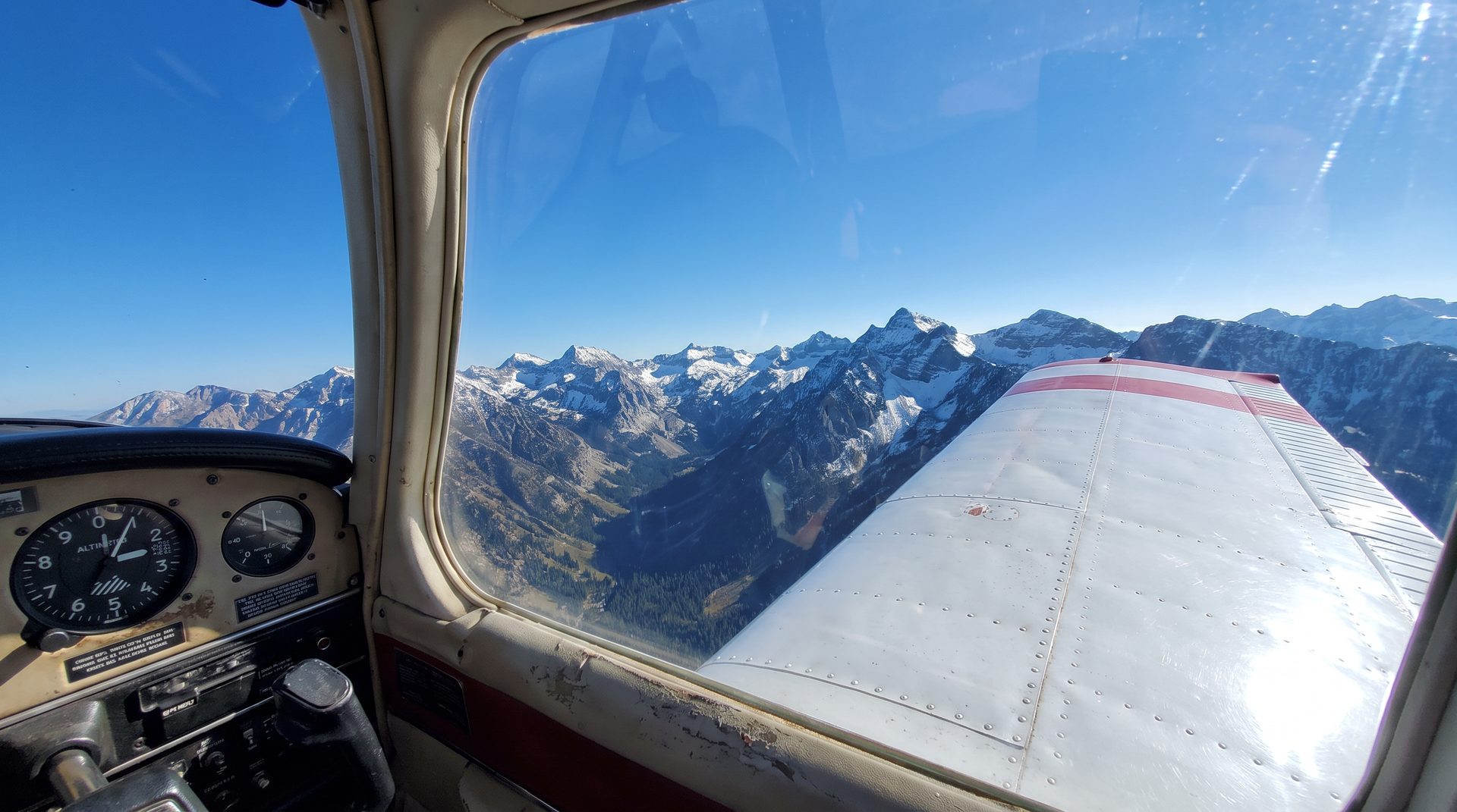 View from a small aircraft cockpit looking out over the wing toward snow-capped Wasatch mountain peaks